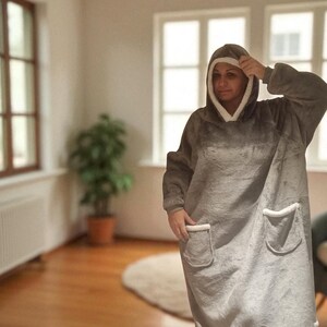 May include: A gray oversized hooded blanket with white trim and two front pockets. The blanket is worn by a person in a well-lit room with hardwood floors and a potted plant.