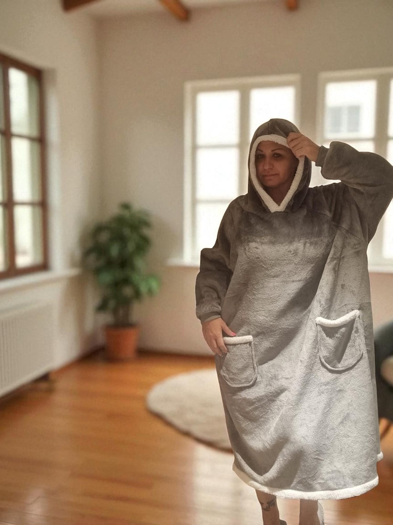 May include: A gray oversized hooded blanket with white trim and two front pockets. The blanket is worn by a person in a well-lit room with hardwood floors and a potted plant.