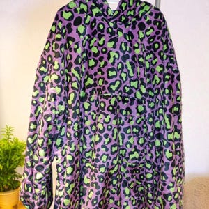 Puede incluir: Una sudadera con capucha morada con un dise&ntilde;o de estampado de leopardo en negro y verde ne&oacute;n. La sudadera de gran tama&ntilde;o est&aacute; colgada en un perchero de metal. La prenda tiene mangas largas y un gran bolsillo delantero.