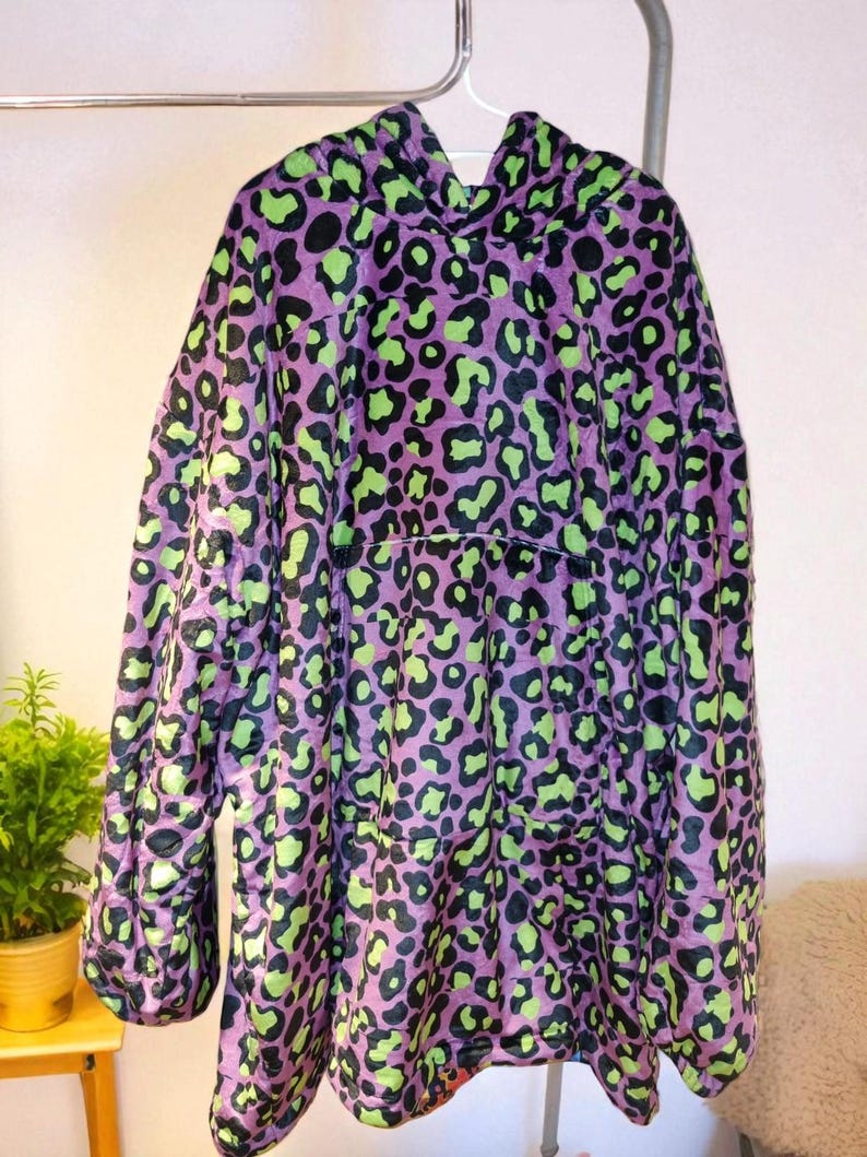 Puede incluir: Una sudadera con capucha morada con un dise&ntilde;o de estampado de leopardo en negro y verde ne&oacute;n. La sudadera de gran tama&ntilde;o est&aacute; colgada en un perchero de metal. La prenda tiene mangas largas y un gran bolsillo delantero.