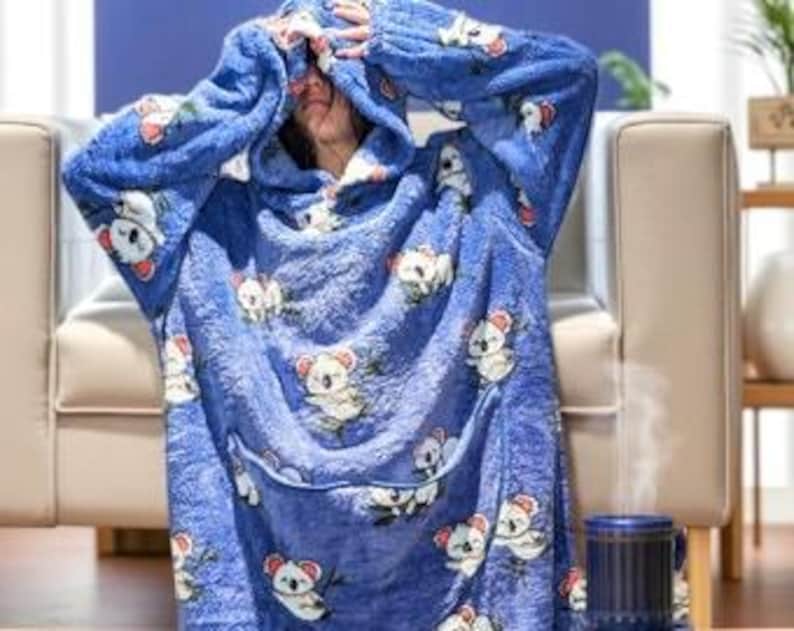 May include: A blue oversized hoodie blanket with a koala bear pattern. The blanket features a hood, a large front pocket, and long sleeves. The koala bears are white with gray accents and are holding branches. The person is wearing the blanket.