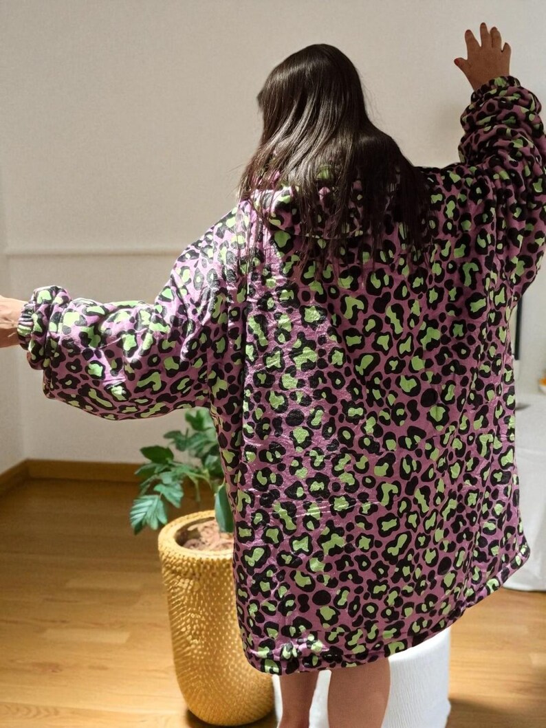 Puede incluir: Una manta con capucha de gran tama&ntilde;o con un estampado de leopardo rosa y verde. La manta tiene mangas largas y un ajuste holgado. La persona que lleva la manta tiene los brazos levantados. El fondo incluye una planta en una maceta amarilla y un suelo de madera.