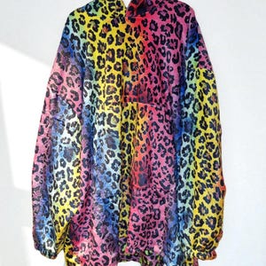 Puede incluir: Chaqueta con capucha con un vibrante estampado de leopardo arco&iacute;ris. La chaqueta presenta un espectro de colores, incluyendo rosa, amarillo, azul y verde, con manchas de leopardo negras. La prenda est&aacute; colgada en una percha blanca sobre un fondo blanco.