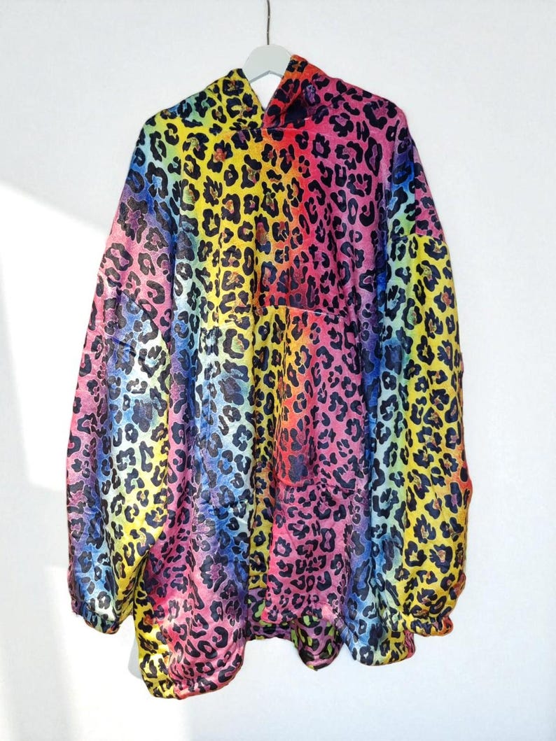 Puede incluir: Chaqueta con capucha con un vibrante estampado de leopardo arco&iacute;ris. La chaqueta presenta un espectro de colores, incluyendo rosa, amarillo, azul y verde, con manchas de leopardo negras. La prenda est&aacute; colgada en una percha blanca sobre un fondo blanco.