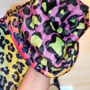 Puede incluir: Una tela con estampado de leopardo colorido en tonos rosa, negro, verde, amarillo y naranja. La tela suave parece estar doblada. El dise&ntilde;o presenta grandes manchas irregulares, creando un patr&oacute;n audaz y llamativo. Probablemente se utiliza para ropa o accesorios.