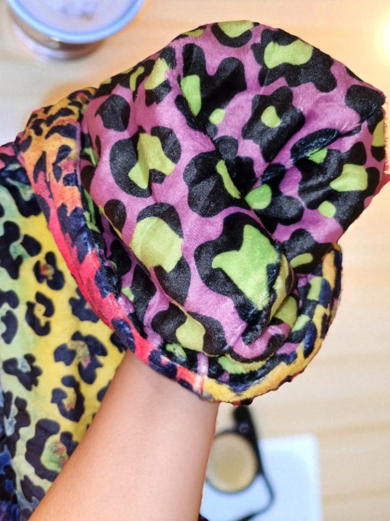 Puede incluir: Una tela con estampado de leopardo colorido en tonos rosa, negro, verde, amarillo y naranja. La tela suave parece estar doblada. El dise&ntilde;o presenta grandes manchas irregulares, creando un patr&oacute;n audaz y llamativo. Probablemente se utiliza para ropa o accesorios.