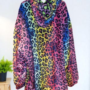 Puede incluir: Una sudadera con capucha con un vibrante estampado de leopardo arco&iacute;ris. El dise&ntilde;o presenta un espectro de colores, incluyendo rosa, amarillo, verde y azul, creando un patr&oacute;n audaz y llamativo. La sudadera est&aacute; colgada en una percha blanca.