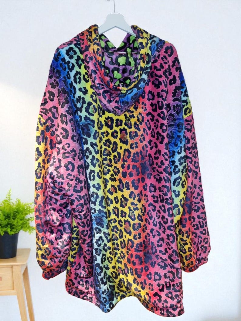 Puede incluir: Una sudadera con capucha con un vibrante estampado de leopardo arco&iacute;ris. El dise&ntilde;o presenta un espectro de colores, incluyendo rosa, amarillo, verde y azul, creando un patr&oacute;n audaz y llamativo. La sudadera est&aacute; colgada en una percha blanca.