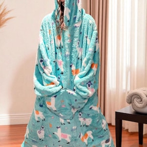 May include: A turquoise hooded blanket with a llama pattern. The blanket is made of a soft, plush material and features a large hood and a front pocket. The llama design includes white, orange, and purple accents.