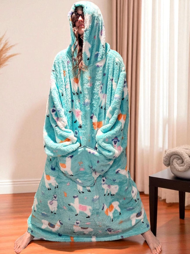 May include: A turquoise hooded blanket with a llama pattern. The blanket is made of a soft, plush material and features a large hood and a front pocket. The llama design includes white, orange, and purple accents.