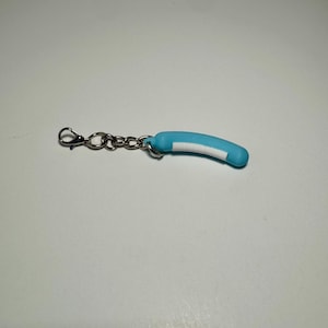 May include: A light blue, curved, cylindrical charm attached to a silver-toned metal chain with a lobster claw clasp. The charm features a smooth, matte finish with a distinct white rectangular stripe running along the inner curve of the object.