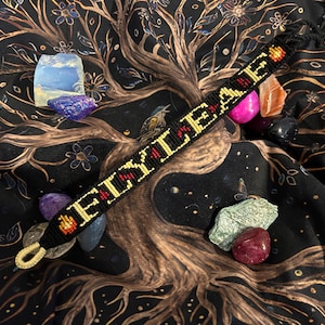 May include: Black friendship bracelet with the word "EILEAH" in yellow letters with red accents. The bracelet is surrounded by colorful crystals and set against a black background with a tree of life design.
