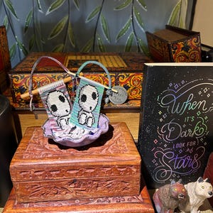 May include: Two small, beaded banners with skull designs are displayed in a decorative bowl. The banners are held by wooden dowels. A wooden box with carved floral designs is in the foreground. A book with the text "When it's Dark Look for Stars" is also visible.