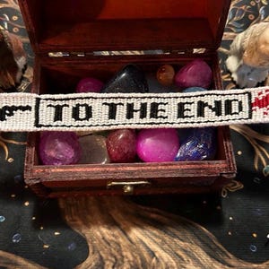 May include: A white woven friendship bracelet with the text "TO THE END" in black letters and a red blood splatter design. The bracelet is displayed on a wooden box filled with colorful stones, with two animal figurines on either side.