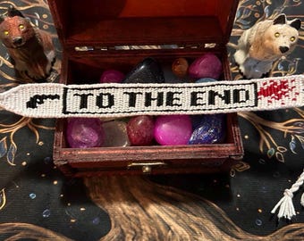 May include: A white woven friendship bracelet with the text "TO THE END" in black letters and a red blood splatter design. The bracelet is displayed on a wooden box filled with colorful stones, with two animal figurines on either side.