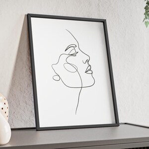 May include: Black framed art print featuring a minimalist line drawing of a woman's profile. The artwork is a continuous line in black against a white background. The frame is rectangular and leans against a wall.