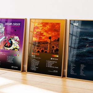 May include: Three framed rectangular posters featuring music album art designs. The left poster is purple with a cartoon character, the middle is orange with a sunflower field, and the right is dark blue with an ocean scene and text.