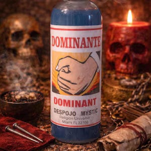 May include: A blue bottle with a white label featuring the word "DOMINANTE" and a drawing of a fist. The bottle is surrounded by a red skull candle, a skull, and other mystical items. The bottle is labeled "DESPOJO MYSTIC".