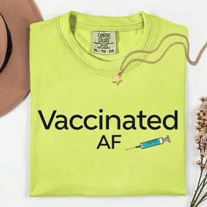 May include: A chartreuse-colored t-shirt with the words "Vaccinated AF" in black, along with a syringe graphic. A gold necklace with a star pendant rests on the shirt. The shirt has a Comfort Colors tag.