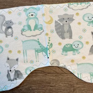 May include: A white baby burp cloth with a whimsical forest animal print. The design features gray squirrels, foxes, and raccoons, along with teal bears, turtles, and deer. The edges are finished with a teal zigzag stitch.