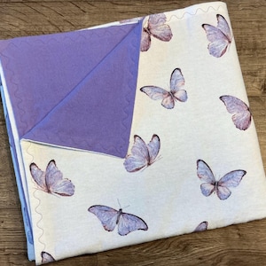 May include: A baby blanket featuring a white background with watercolor-style purple butterflies. The blanket has a lavender interior and a decorative stitched edge. The blanket is folded, showcasing the contrasting colors and patterns.