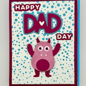 May include: A handmade Father's Day card with a blue and burgundy border. The card features the words "HAPPY DAD DAY" in burgundy and blue, with a cute pink monster illustration. The background has a dotted pattern.