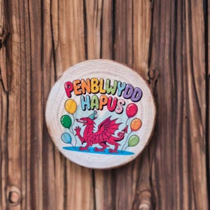 Puede incluir: Rodaja de madera redonda con una ilustración colorida. La ilustración presenta un dragón rojo con un gorro de fiesta, rodeado de globos. El texto "PENBLWYDD HAPUS" está escrito en arcoíris. El fondo es de madera.