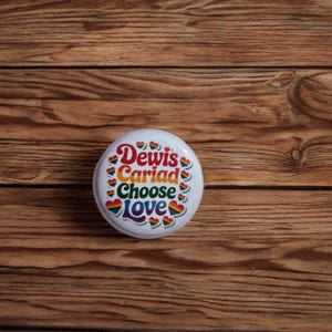 Puede incluir: Un botón blanco con las palabras "Dewis Cariad Choose Love" en colores arcoíris. El botón tiene diseños de corazones arcoíris y está sobre un fondo de madera.