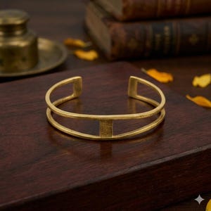 Può includere: Un bracciale a polsino color oro con un design centrale rettangolare. Il bracciale ha un design a due barre ed è esposto su una superficie di legno scuro. Il gioiello è un accessorio semplice ed elegante.