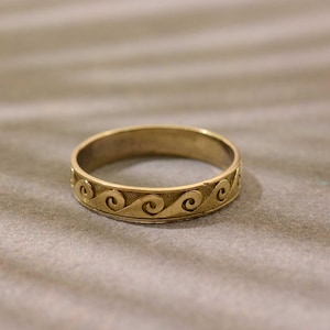 Może przedstawiać: Złoty pierścionek z powtarzającym się wzorem fal wokół obrączki. Pierścionek wykonany jest z błyszczącego metalu i leży na neutralnej powierzchni. Wzór pierścionka przedstawia serię stylizowanych fal.