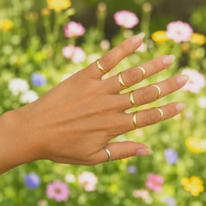 Op de afbeelding: Een set van zes goudkleurige metalen ringen gedragen aan de vingers. Vijf ringen zijn geplaatst op de bovenste vingerkootjes, terwijl één zilverkleurige ring aan de basis van de duim wordt gedragen. De ringen hebben een glad, effen ontwerp.