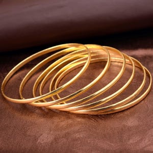 May include: A collection of gold bangles, each with a smooth, polished surface. The bangles are stacked together, creating a layered effect. The warm gold color contrasts with the brown background.