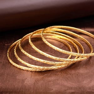 May include: Six gold bangles with intricate floral patterns are arranged on a brown surface. The bangles are stacked together, showcasing their detailed design and warm, metallic color. The background is a darker shade of brown.