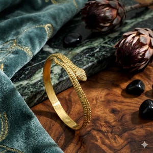 May include: A gold-colored snake bangle bracelet. The bracelet features a textured design and a snake head detail. The jewelry is displayed on a wooden surface with decorative elements, including a green velvet cloth and black stones.