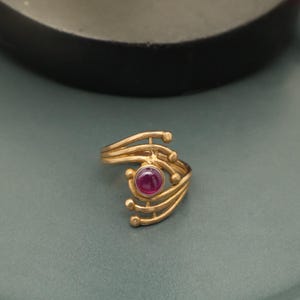 Peut inclure: Une bague dorée avec une pierre précieuse rouge ronde au centre. La bague présente un design abstrait avec des lignes courbes et de petits accents ronds. La bague est posée sur un fond vert.