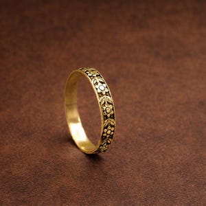May include: A gold-colored ring with a floral pattern. The ring features a repeating design of flowers and leaves around the band. The ring is set against a brown background.