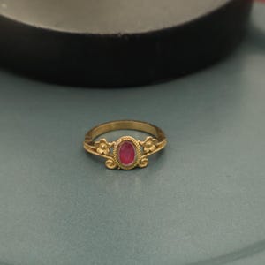 May include: A gold ring with a central oval-shaped red gemstone. The ring features floral accents on either side of the gemstone. The ring is set against a teal background.