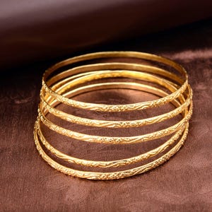 May include: A stack of six gold bangles with intricate floral and swirl patterns. The bangles are displayed on a brown surface, and the light reflects off the polished metal. The bangles are a classic jewelry item.