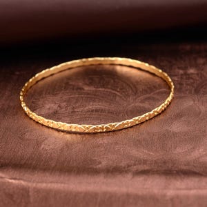 May include: A gold-colored bangle bracelet with a textured, repeating pattern. The bracelet is circular and rests on a brown surface. The bangle is a jewelry item.