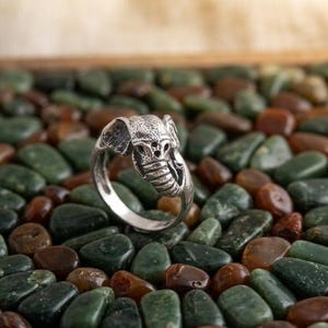 May include: A silver ring with an elephant head design. The ring is displayed on a bed of green and brown stones. The elephant's face has detailed, textured features.
