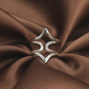 Puede incluir: Un anillo de tono plateado con un diseño geométrico calado que presenta dos bandas simétricas en forma de punta de flecha que se unen en el centro, creando una silueta angular y hueca con un acabado metálico pulido y reflectante.