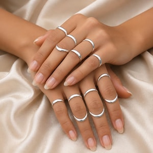 Può includere: Un set di dieci anelli color argento indossati sulle dita. Gli anelli presentano fasce lisce e semplici, alcune progettate come mezzelune aperte che avvolgono le articolazioni. Il metallo ha una finitura lucida e riflettente su tutto il set.