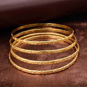 May include: A stack of five gold bangles. Each bangle has a textured, engraved design. The bangles are displayed on a brown surface. The gold color of the bangles contrasts with the dark background.
