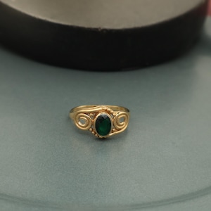 Puede incluir: Un anillo de oro con una piedra preciosa ovalada de color verde oscuro en el centro. El anillo presenta un diseño decorativo en espiral a cada lado de la piedra preciosa. El anillo está sobre un fondo verde apagado.