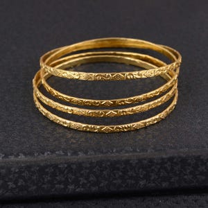 May include: Four gold-colored bangles with intricate, repeating patterns. The bangles are stacked on top of each other, with the top bangle slightly out of focus. The background is a dark, textured surface.