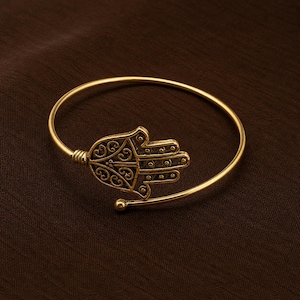Könnte beinhalten: Ein goldfarbenes Hamsa-Hand-Armband. Das offene Handdesign ist mit komplizierten Mustern versehen und an einem einfachen, gebogenen Band befestigt. Das Armband hat eine kleine Kugel am Ende des Bandes. Der Hintergrund ist einfarbig braun.