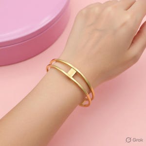 May include: A gold-colored bracelet with a geometric design. The bracelet features two parallel bands connected by a small square in the center. The jewelry is worn on a person's wrist.