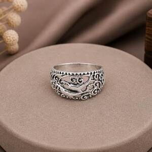 May include: A silver ring with an openwork design featuring a stylized wave pattern. The ring is wide and sits on a round, neutral-toned surface. The background includes a brown fabric and a wooden box.