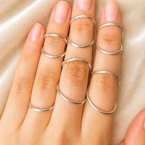 May include: A collection of seven thin, smooth, polished silver-tone metal rings worn on the fingers. Each ring features a simple, minimalist circular band design with a reflective metallic finish, distributed across the fingers of the hand.