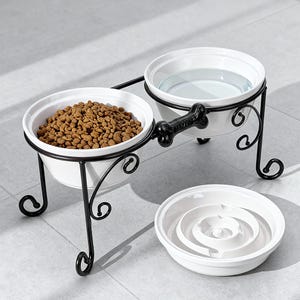 May include: A white ceramic pet bowl set with a black metal stand. One bowl contains dry pet food, the other water. A separate white slow feeder bowl is also present. The stand has a bone-shaped handle.